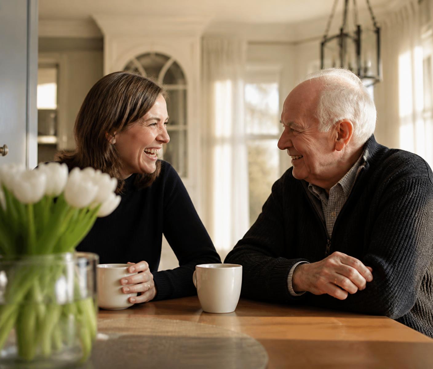 An adult daughter laughing with her elderly father over coffee at a sunlit kitchen table
